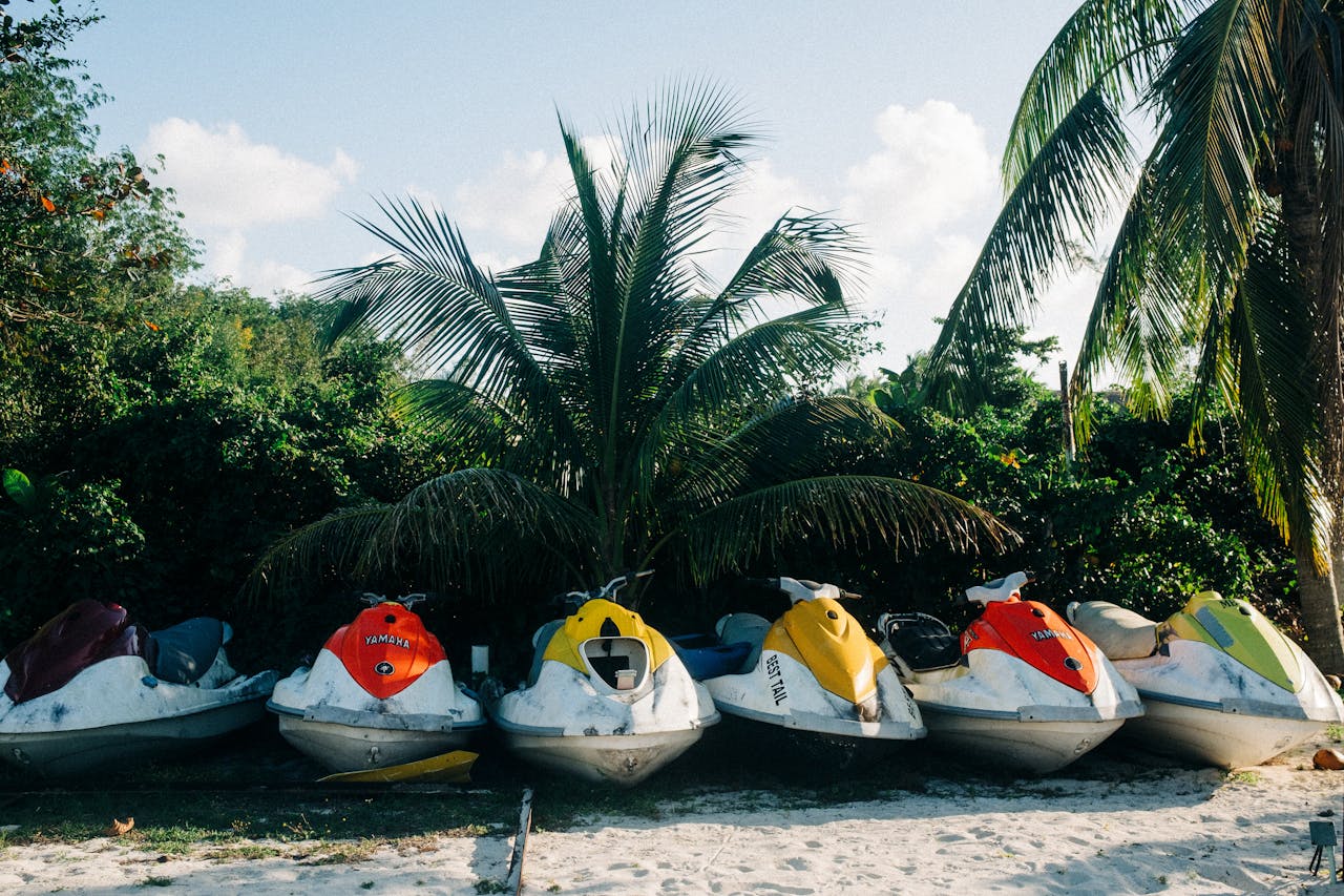 Vibrant jet skis parked on a sandy tropical beach with lush palm trees and clear blue skies.